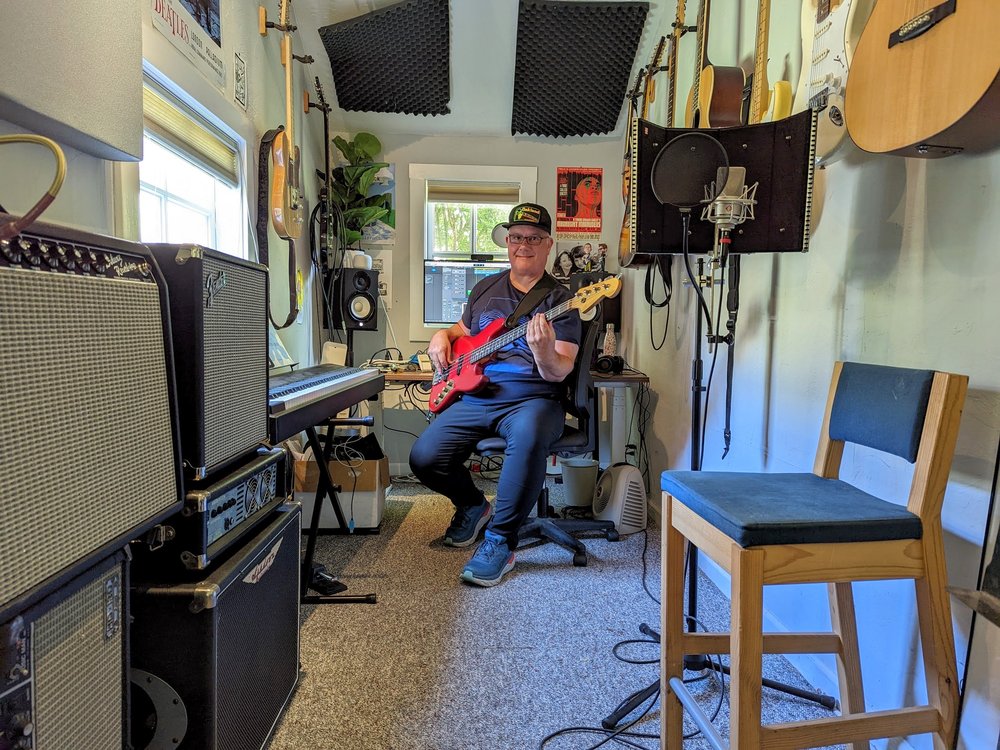 Ben in his home recording studio. He’s sitting and holding a red bass guitar with various instruments and audio equipment around him.