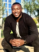 Chudi Iregbulen sitting with hands folded, leaning towards the camera smiling and wearing a black jacket