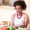 Black Foodie founder Eden Hagos smiles in a white halter top with red necklace, hoop earrings and upswept hair, as she chops green veggies at the kitchen counter.