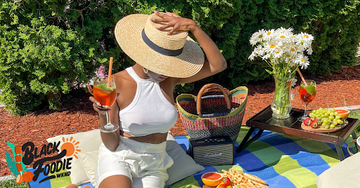 A woman wearing white halter top, shorts and straw hat holds a pink fruity drink while seated on a blue-and-green striped blanket in a backyard picnic.