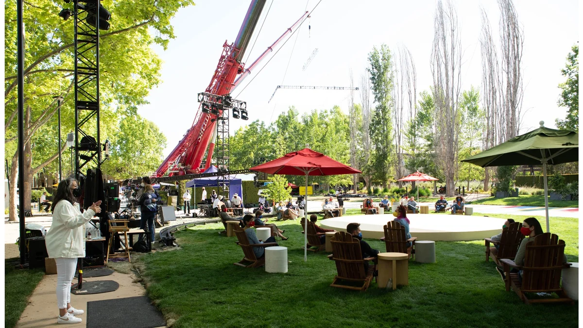 Photo showing the “quad” at Google’s Mountain View campus. People wearing masks are sitting in chairs on the lawn around a circular stage; a large crane is visible in the background.