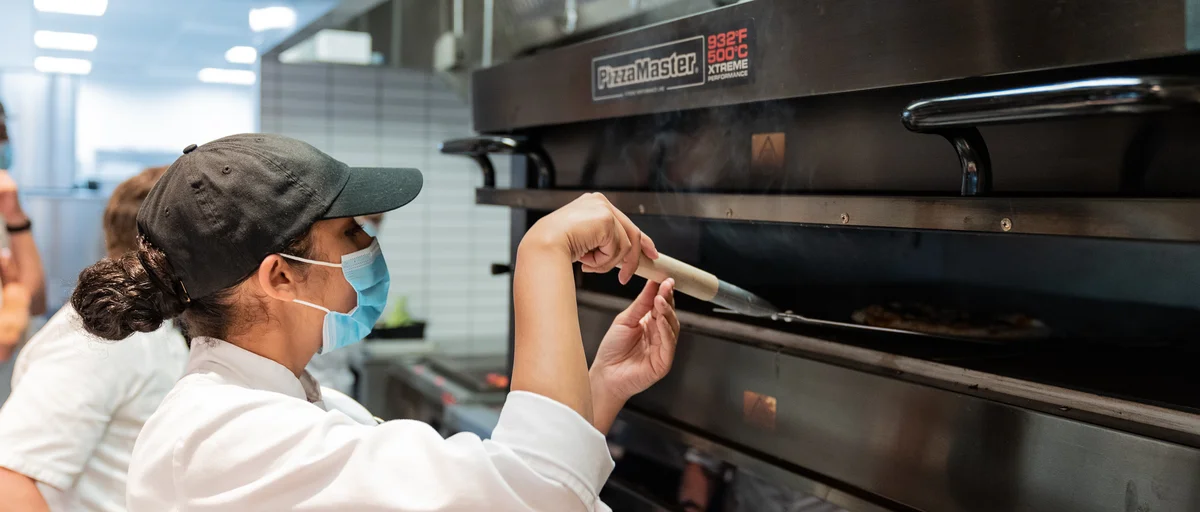 A cook uses a pizza tray to guide a pizza into an oven.