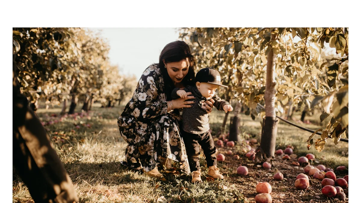 Cheryl is in a Black, flowery dress, with dark hair and fair skin. She is smiling and crouched down holding her son under his arms while he is standing looking down. Her son is a toddler in a gray shirt, black pants, brown shoes, with a black baseball hat