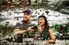 A bearded man in a blue baseball cap and a woman with long black hair in black cat-eye glasses take a photo of themselves in front of an artistic hall of mirrors surrounded by mirrored orbs.