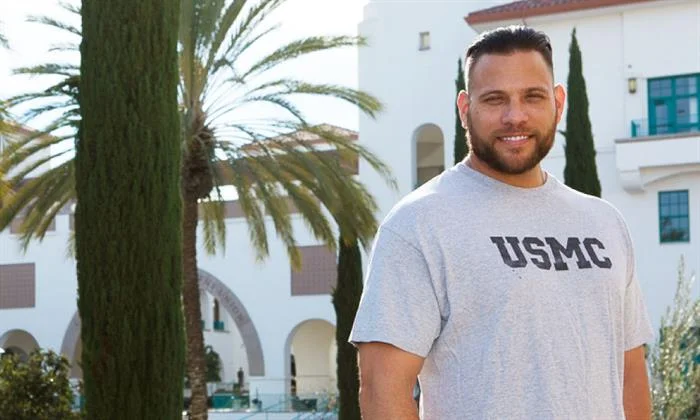 A man wearing a gray shirt with "USMC" in blue letters looks at the camera, in front of a Spanish-style building.