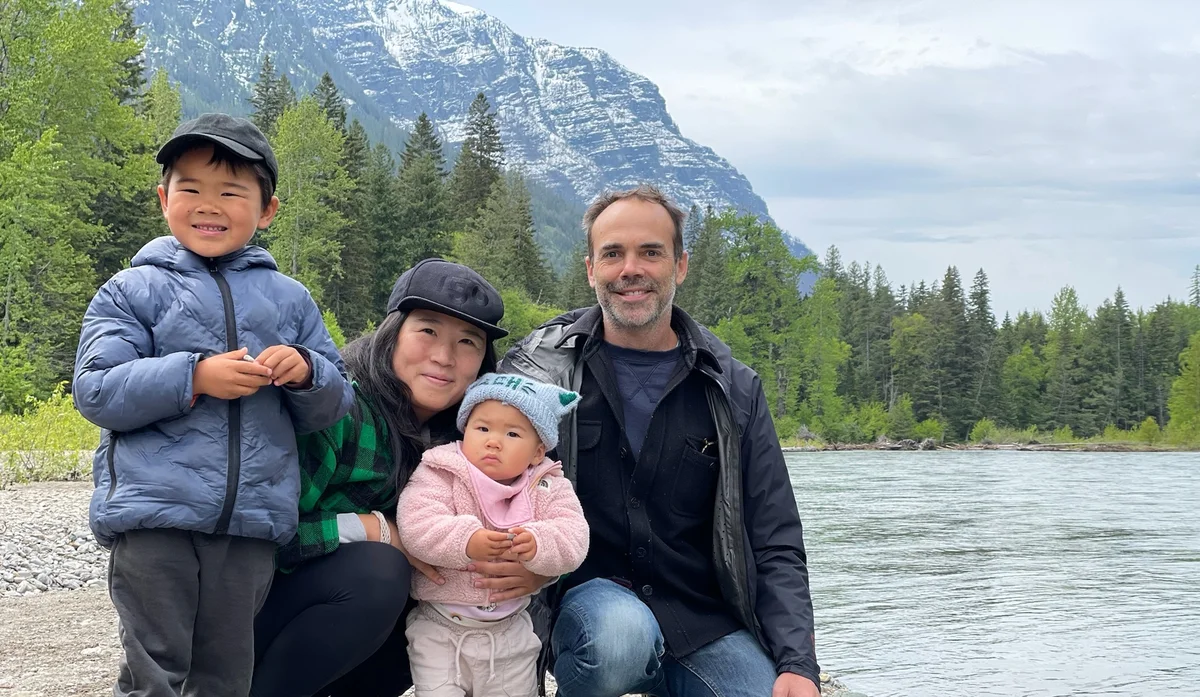 An image of a man, woman, and two children kneeling by a lake with large mountains in the background.