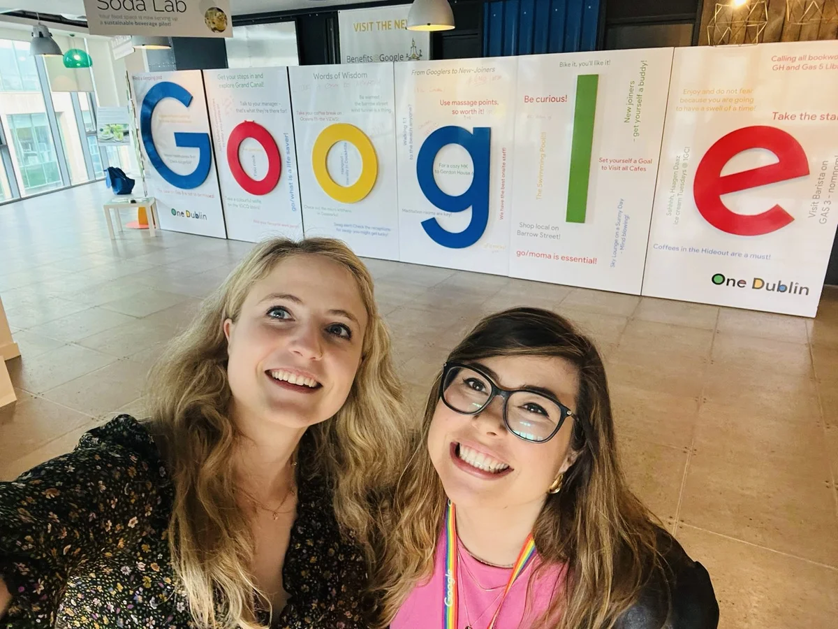 Desi und ihre Kollegin Kathi machen ein Selfie vor einem Google-Schriftzug im Dubliner Google-Büro und lächeln dabei in die Kamera.