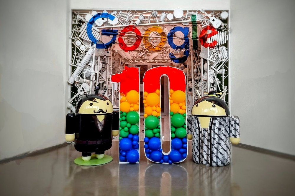 An image shows a section of the Google Indonesia office. In the image, there is a physical “10” logo, indicating Google Indonesia’s 10th Anniversary, with two physical Android mascots standing on each side. There is a Google logo above.