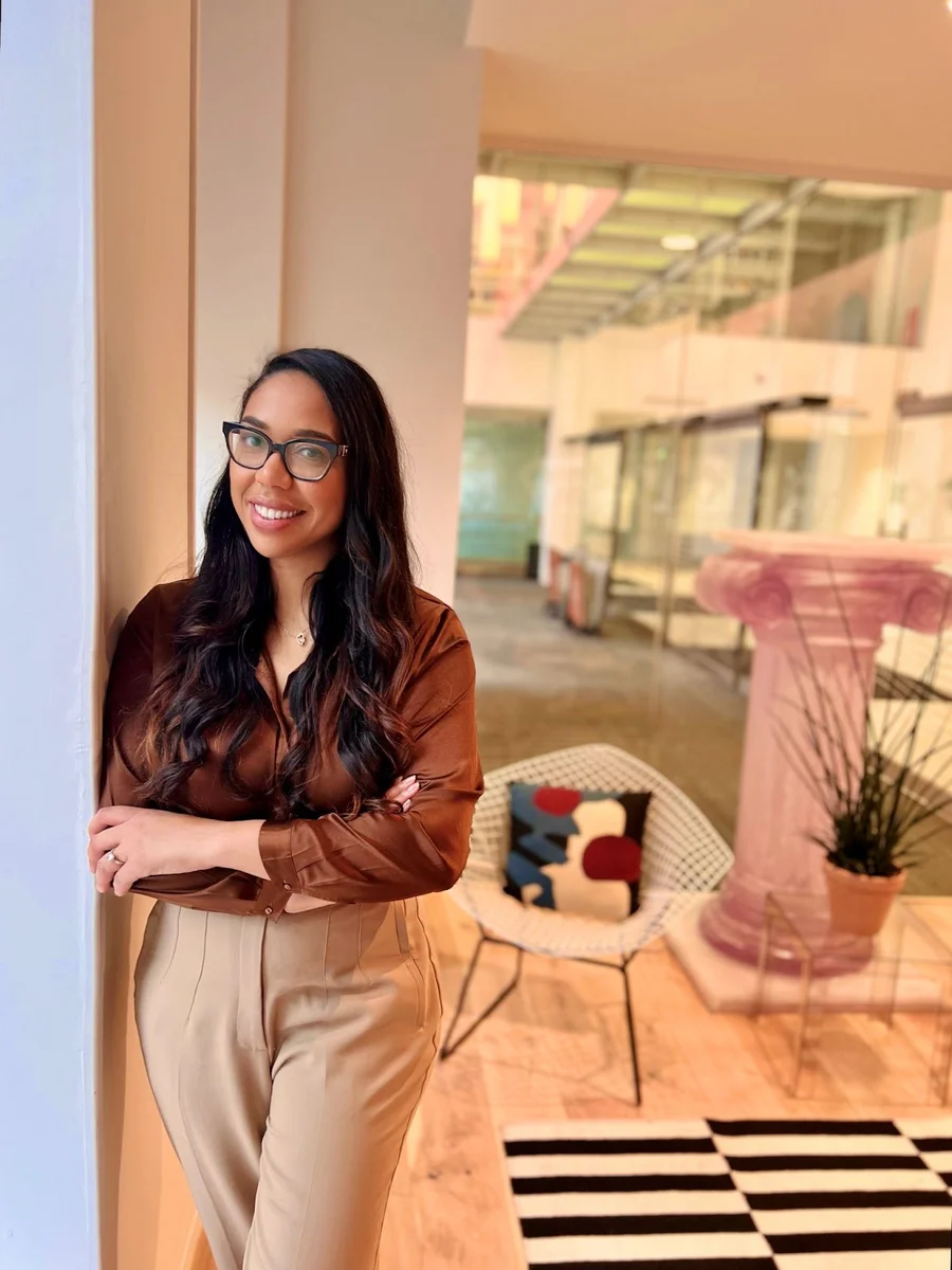 A woman with long dark hair and glasses leans against the wall of a brightly-light room, with her arms folded across her torso. She's wearing a burgundy top and taupe pants, and there's a striped rug and a gray chair visible in the background.