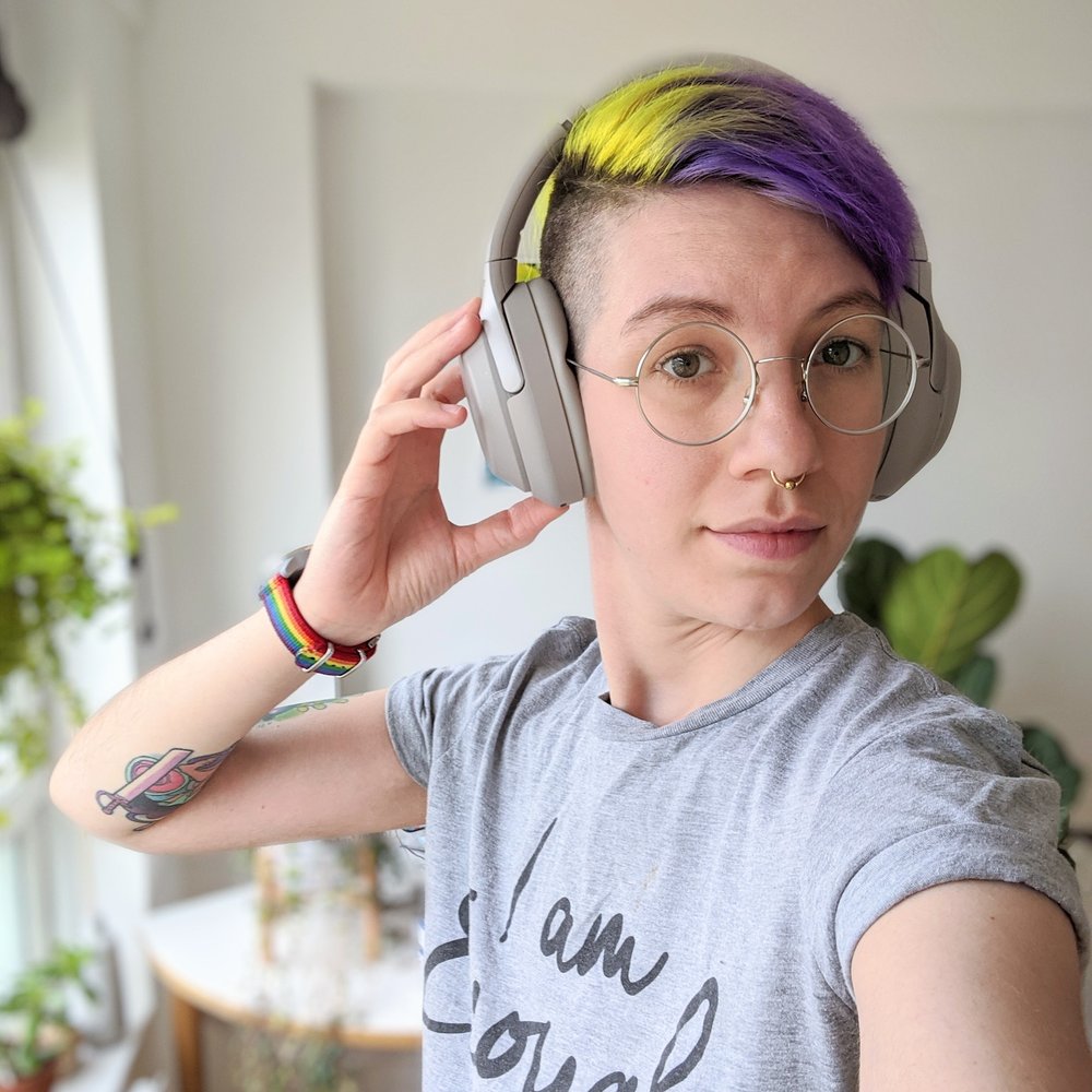 Androgynous thin white person with short hair dyed purple and yellow. They wear noise cancelling headphones and a grey t-shirt that says “I am enough”. Lio also wears round glasses and a watch with a rainbow strap. Their right hand is reaching up to their headphones, revealing a Steven Universe tattoo. They look proud and optimistic. In the background are plants and sunlight shines through a window.