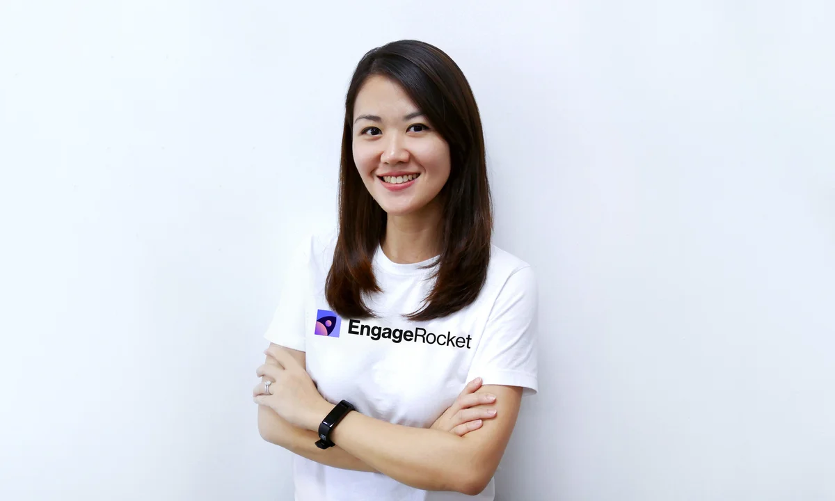 Picture of Dorothy Yiu, the founder of EngageRocket, wearing a white t-shirt with the company logo, against a white background. She is looking at the camera and smiling with her arms crossed.