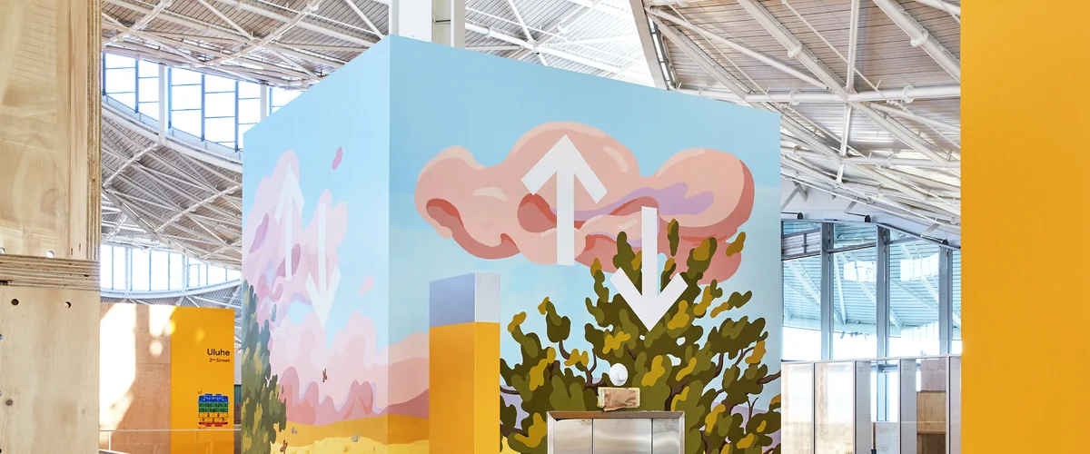 A photograph of the top of an elevator shaft in a large, open building. The elevator is painted with a mural that features illustrated pink clouds and a green tree.