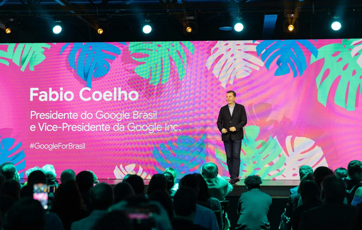 Fabio Coelho, a white man wearing a black suit in onstage in front of an audience. In the back, there’s a screen with a pink background and his name and occupation title.