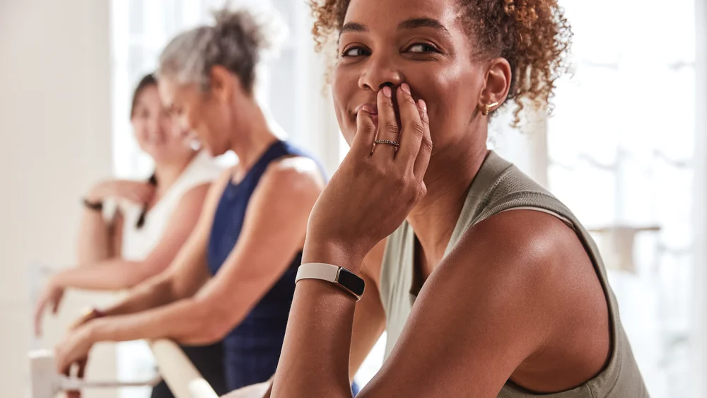 A woman wearing a Fitbit Luxe tracker leaning on a ballet barre with her fingers touching her face; two other women, out of focus, are also leaning on the barre in the background.