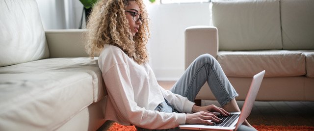 La imagen muestra a una mujer joven con cabello rizado y anteojos sentada en el suelo con una computadora portátil en su regazo, escribiendo en el teclado.