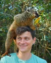 Matt smiles, wearing a light blue t-shirt and looking into the distance. A lemur is perched on his head, holding a piece of fruit and, looking in the opposite direction.