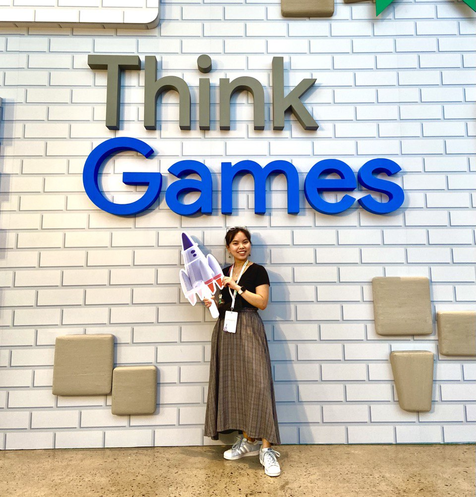 Gia, wearing a black t-shirt and patterned skirt, stands in front of a white brick wall with the Think Games logo and holds a cardboard cut-out of a rocket ship.