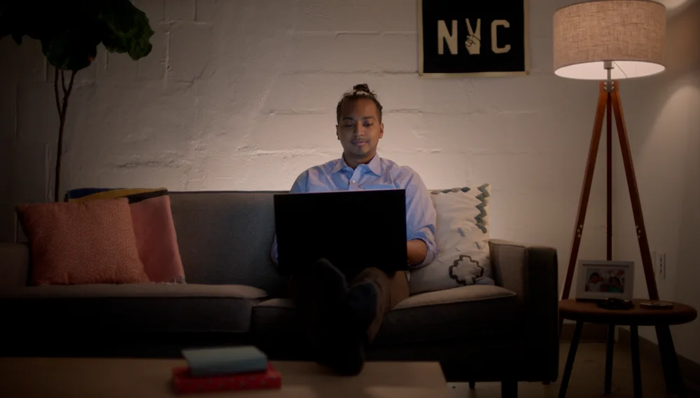 A young man is sitting on his couch with a laptop.