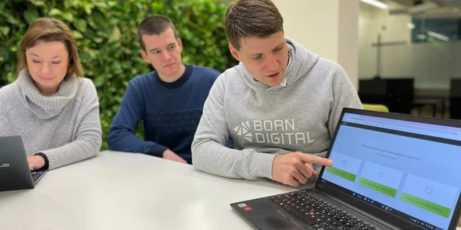 Three people sit around a white table, looking an open laptop screen with the Born Digital website open. The man nearest the computer wears a gray sweatshirt that has "Born Digital" written in white letters.