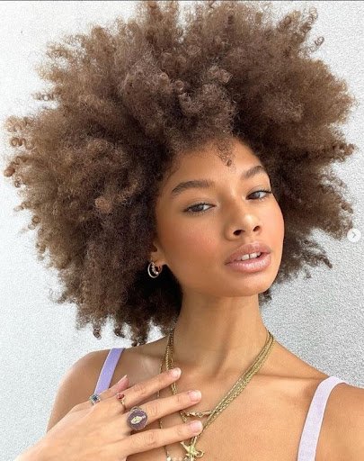 Hannah smiles as she shows off her brown curly hair, which she wears in an afro. She has on a tank top, gold necklaces, hoop earrings and rings on her fingers.