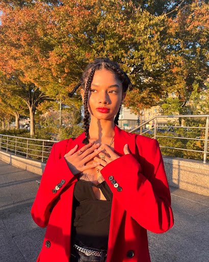 Hannah stands on a sidewalk in Manhattan, New York, with trees and a metal fence behind her. She wears a red, oversized blazer with a lacy black top, red lipstick and her hair in braids.