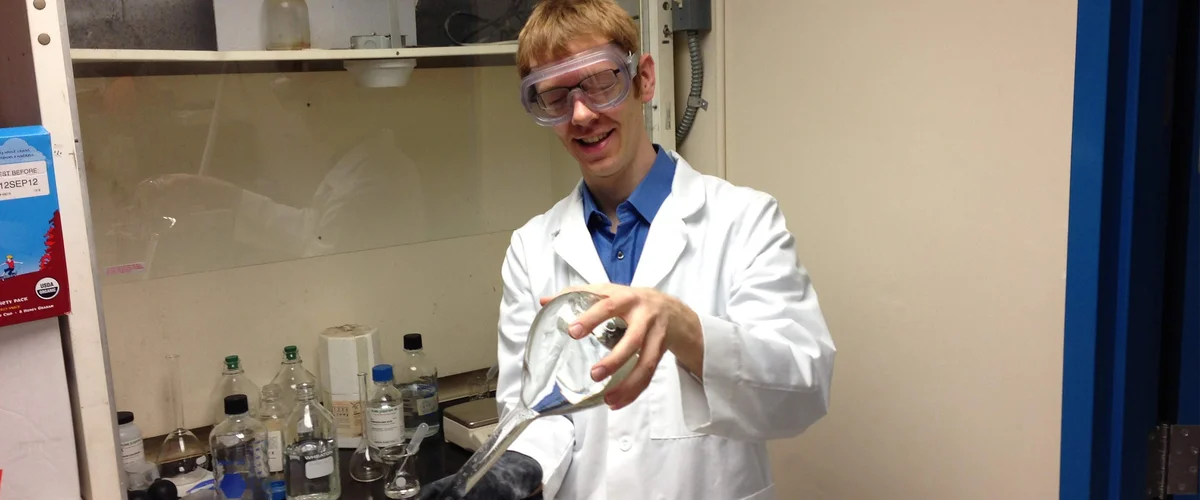 Garrett wearing a lab coat and protective goggles holds a glass flask while standing in front of a lab counter and bottles.
