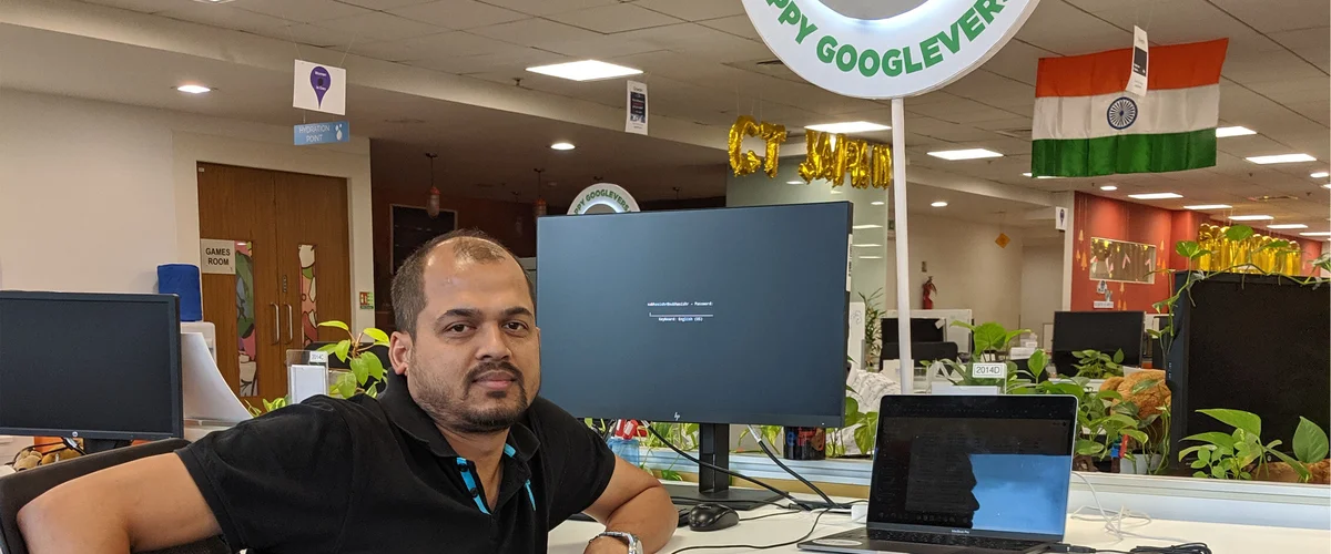 Subhasish sits at his desk at the Hyderabad office wearing a black shirt. On the desk is a laptop and a monitor. In the background are other desks and computers and decorations like  office plants, an Indian flag, and the bottom of a sign that says Happy