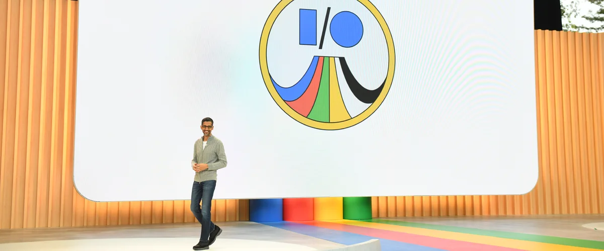 Sundar Pichai at the front left of a large stage with a colorful I/O logo behind him.
