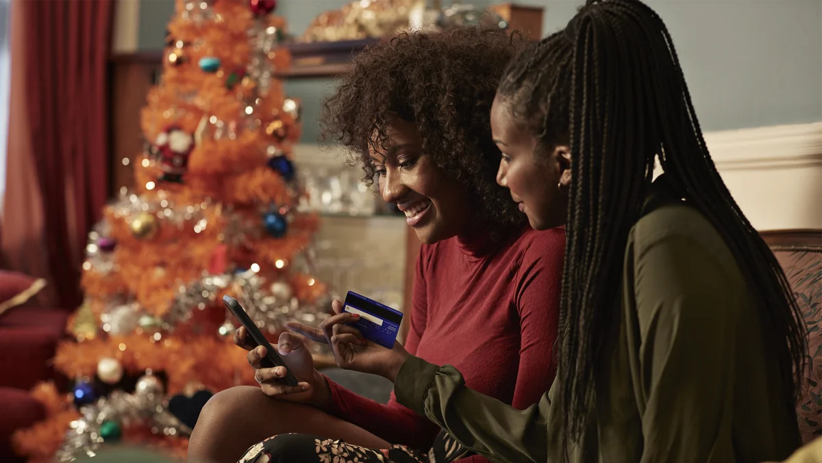 Two Black women sit on their couch at home online shopping together. One woman holds her phone while the other woman looks on and holds her credit card. There is an artificial orange Christmas tree in the background.