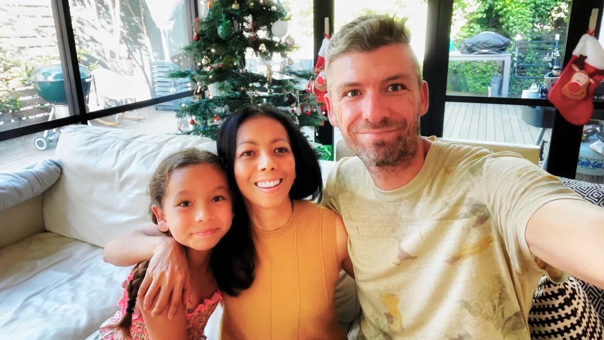 Patricia Torres, her husband Chris and daughter Natasha are smiling and sitting on a couch with their arms around each other. In the background is a Christmas tree.