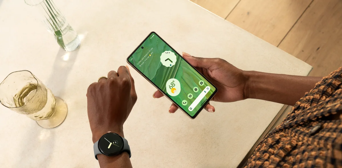 Eine Person hält ein Pixel 7 Smartphone in der rechten Hand, auf dem der Homescreen zu sehen ist. Am linken Arm trägt die Person eine Pixel Watch.