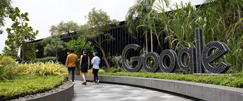 A Google logo sign along a path surrounded by plants and trees. A small group of people are talking and walking together in the background.