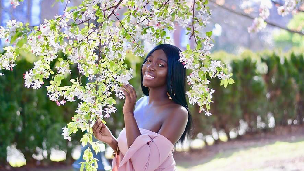 Nyamekye Nti is standing outside, holding tree branches with pink flowers. She is smiling at the camera and wearing a pink, off-the-shoulder top.