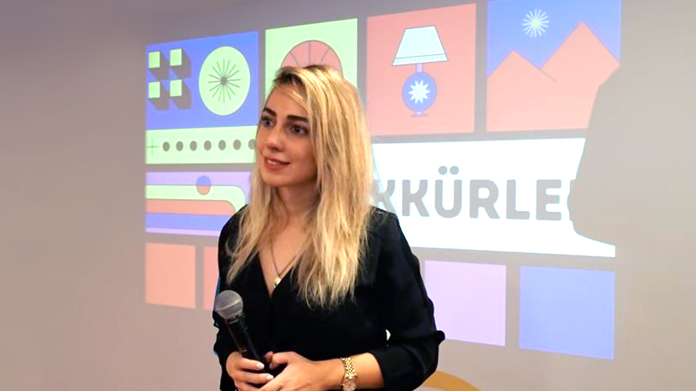Photo of Merve Isler standing in front of a screen. She's holding a microphone and looking into an audience.