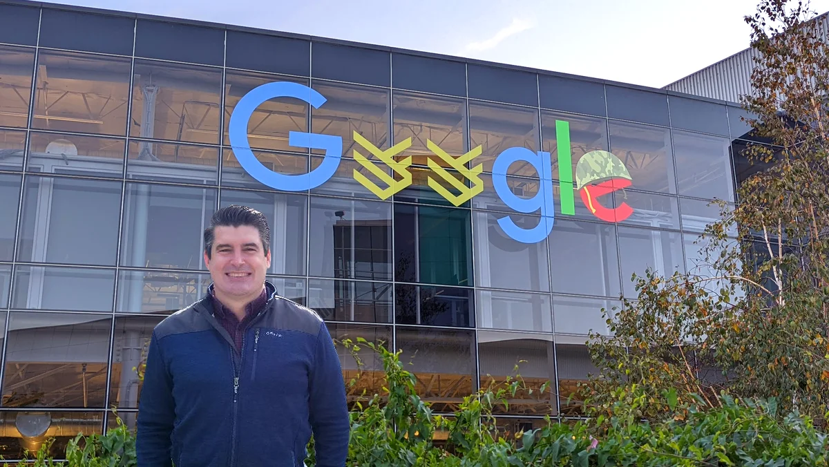 Michael está de pie frente a un edificio de cristal, vestido con una chaqueta azul sobre una camisa de botones de color púrpura oscuro. Detrás de él está el logotipo de Google modificado al logotipo de Google Veterans Network.