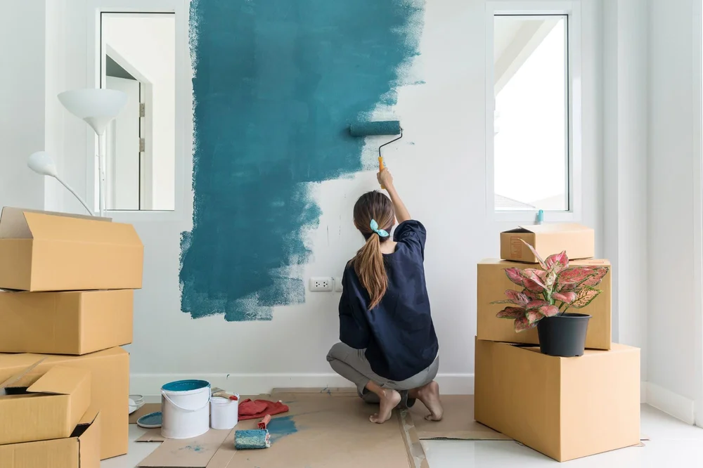 A woman is painting a blank white wall blue, surrounded by moving boxes and a plant