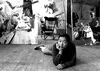 A black-and-white photograph of painter Bernice Bing lying on her stomach on the floor of her studio, her head propped on her hand and looking directly at the camera. In the background, you can see three of her paintings and a wood rocking chair.