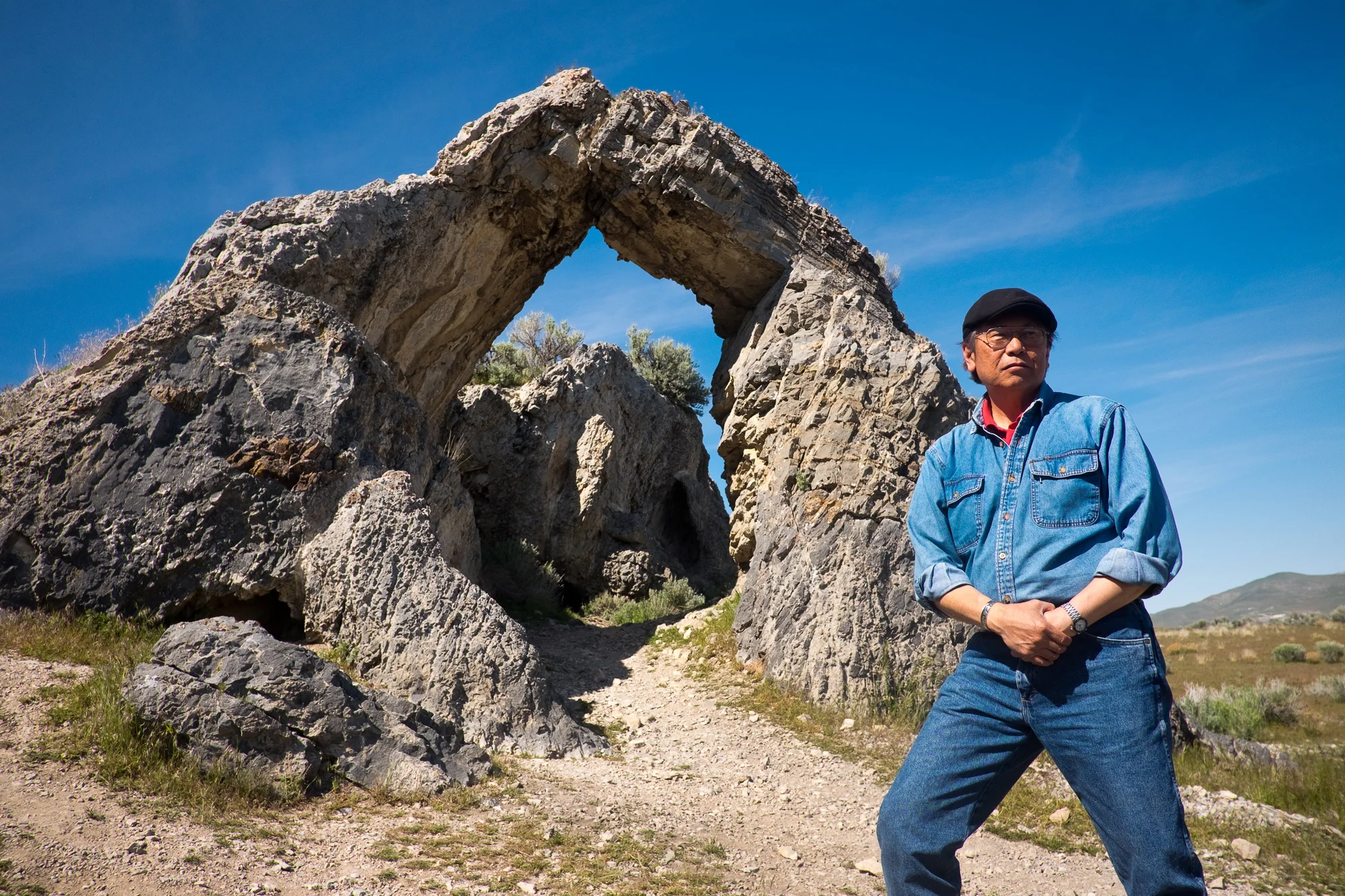 A digital color photograph of Chinese-American photographer Corky Lee, standing in the foreground in a natural outdoors landscape wearing a light denim shirt and dark blue jeans. In the background are natural stone archies.