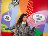 A woman wearing a black and white sweater poses in front of a colorful wall with bright red, blue, purple and yellow. She’s pointing to two speech bubbles that say “Ruth Asawa!” and “Carlos Villa!”