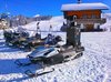 A row of snowmobiles, one of which with a camera attached, sit at the base of a ski slope.