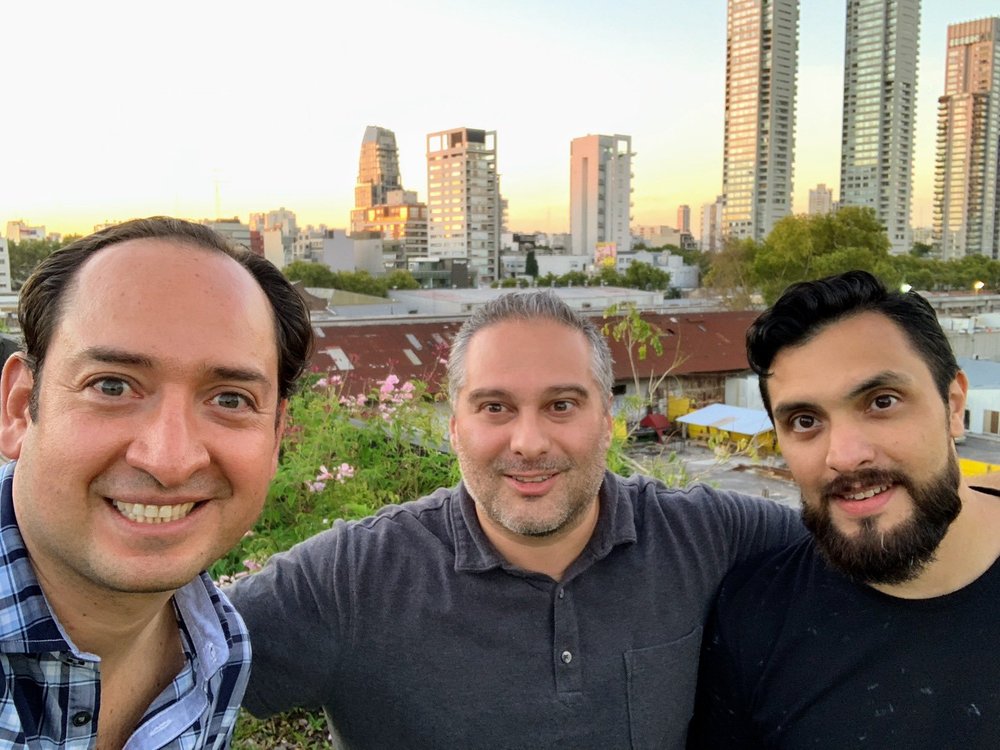 Three men smile at the camera with a city skyline backdrop behind them.