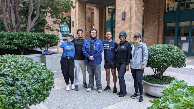 Six people standing in front of a brick building, next to a variety of plants. They are all smiling at the camera, one person is holding up a peace sign.