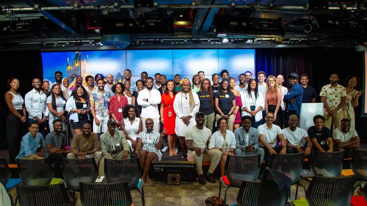 Approximately 40 Black Founders Fund recipients and Googlers from Europe & Africa are gathered in an auditorium—most are standing, while a row sit on the edge of the stage. Behind them is a blue screen projecting an image of the Tower of London.