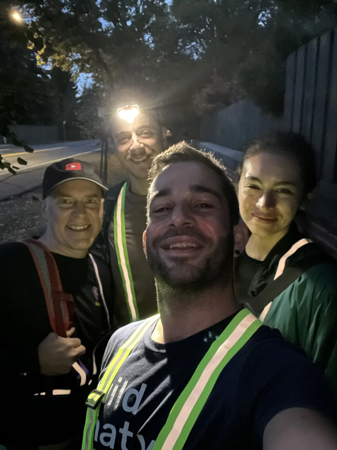 Selfie of four people in the evening. They are all smiling at the camera, and one person is wearing a headlight.