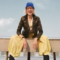 There is an older woman in a leather jacket and a beanie sitting on a bench smiling while wearing Inspire 3.