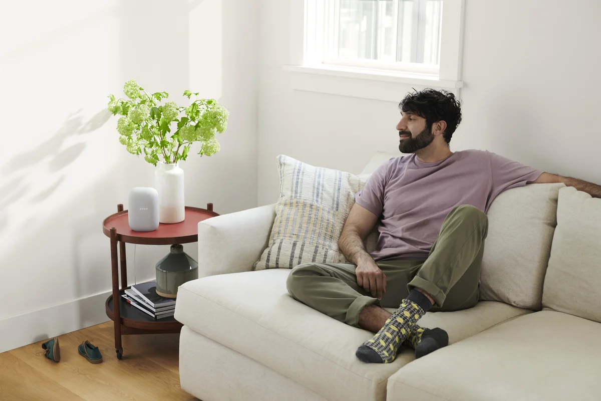 Image shows a man sitting on a white couch in a living room next to a window. On the side table there is a potted plant as well as a Nest Audio.