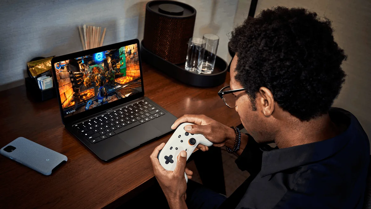 A young man dressed in black sits at a brown desk with a white controller playing Stadia games on his black laptop.