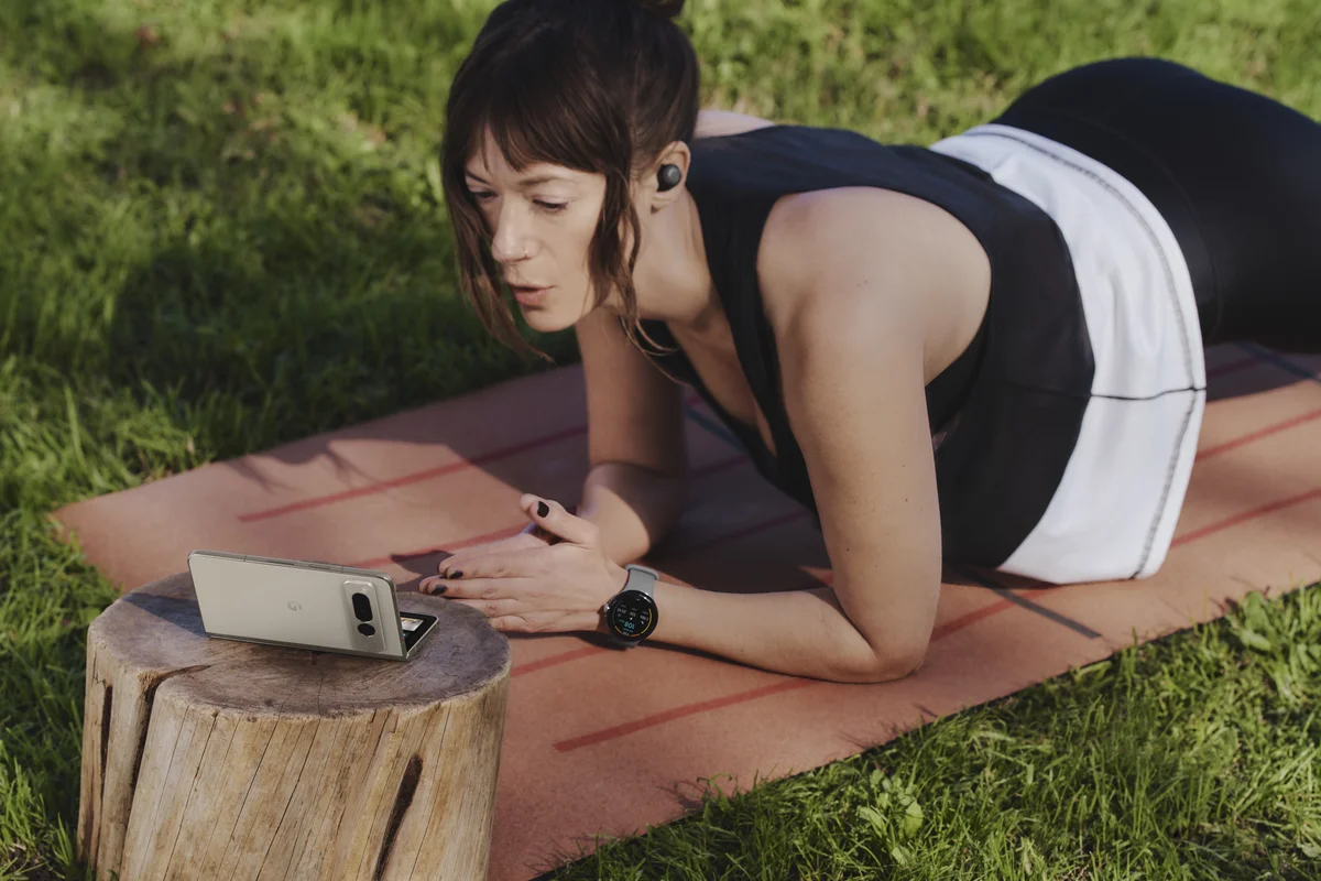 Photo of a woman holding a plank on an orange yoga mat on top of a grassy lawn. She’s wearing Pixel Buds and looking at a Pixel Fold, which is sitting in front of her on top of a stump. It’s open in tabletop mode.