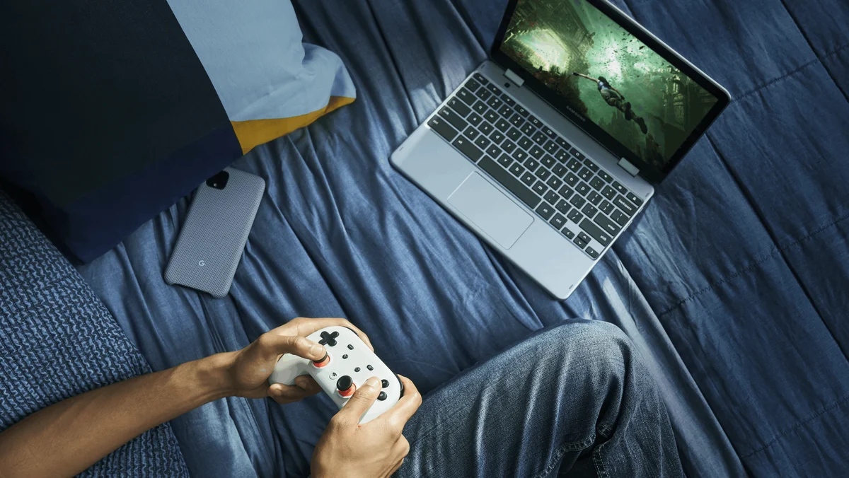 A blue image shows a top-down perspective of a person's hands holding a white controller while sitting on a blue bed and playing Stadia games on a silver laptop.