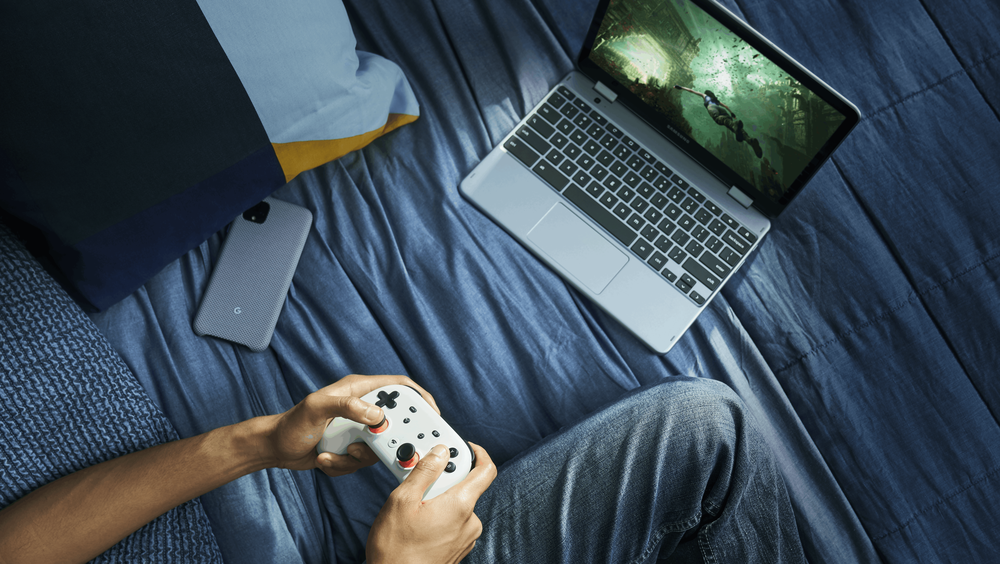 Sitting on a blue bed, a player holds a white Stadia controller and games on their laptop.